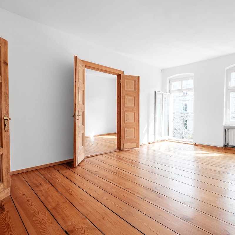 Grande pièce vide avec parquet en bois clair, murs blancs, deux portes doubles en bois ouvertes et fenêtres donnant sur un balcon.
