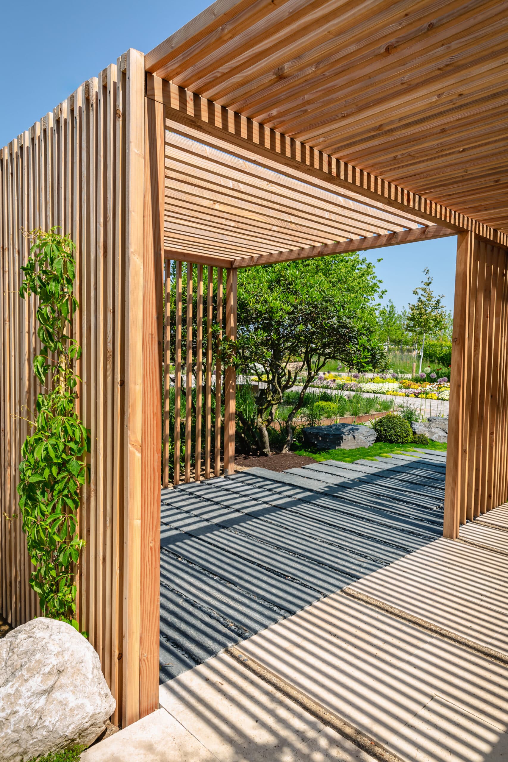 Structure en bois moderne avec lattes verticales et horizontales, projetant des ombres rayées sur un chemin paysager gris et beige. Une plante verte y grimpe.