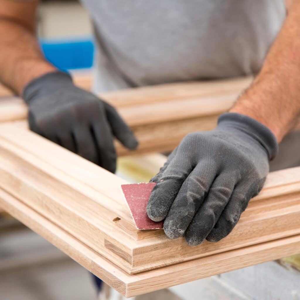 Main gantée ponçant un cadre en bois clair avec du papier de verre rouge. Un menuisier travaille minutieusement.