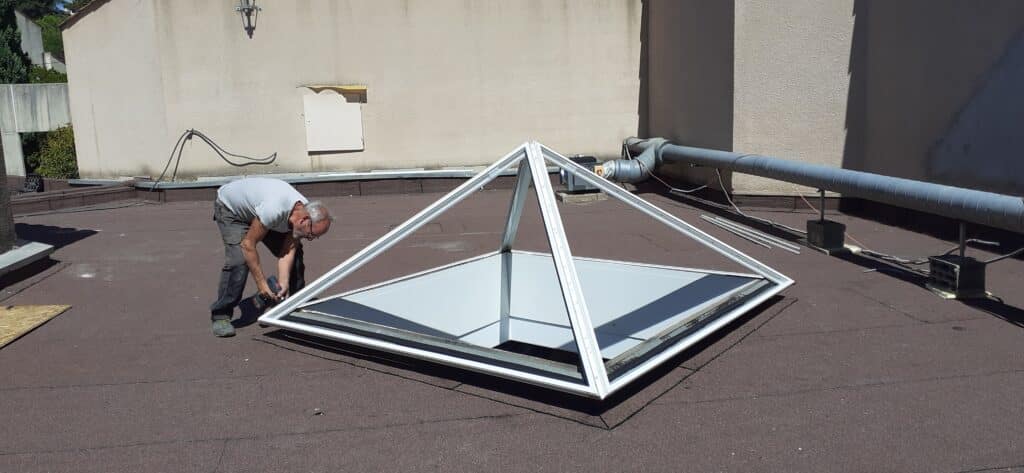 Ouvrier installant un lanterneau pyramidale blanc sur un toit plat goudronné par une journée ensoleillée.