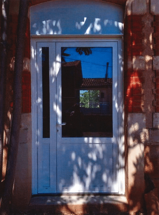 Porte d'entrée blanche moderne vitrée avec imposte, installée dans une façade en briques rouges. Reflet bleu du ciel et ombres d'arbres.