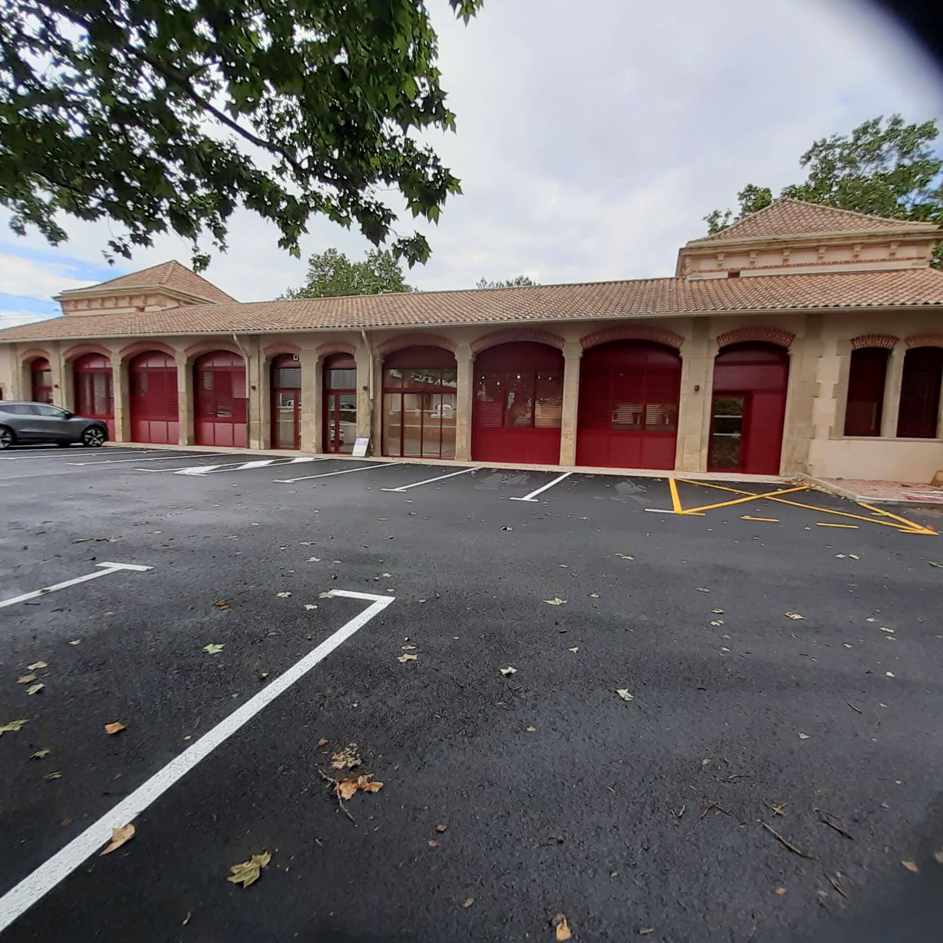 Façade de bâtiment aux arcades et volets rouges sous un toit en terre cuite avec un parking goudronné au premier plan.