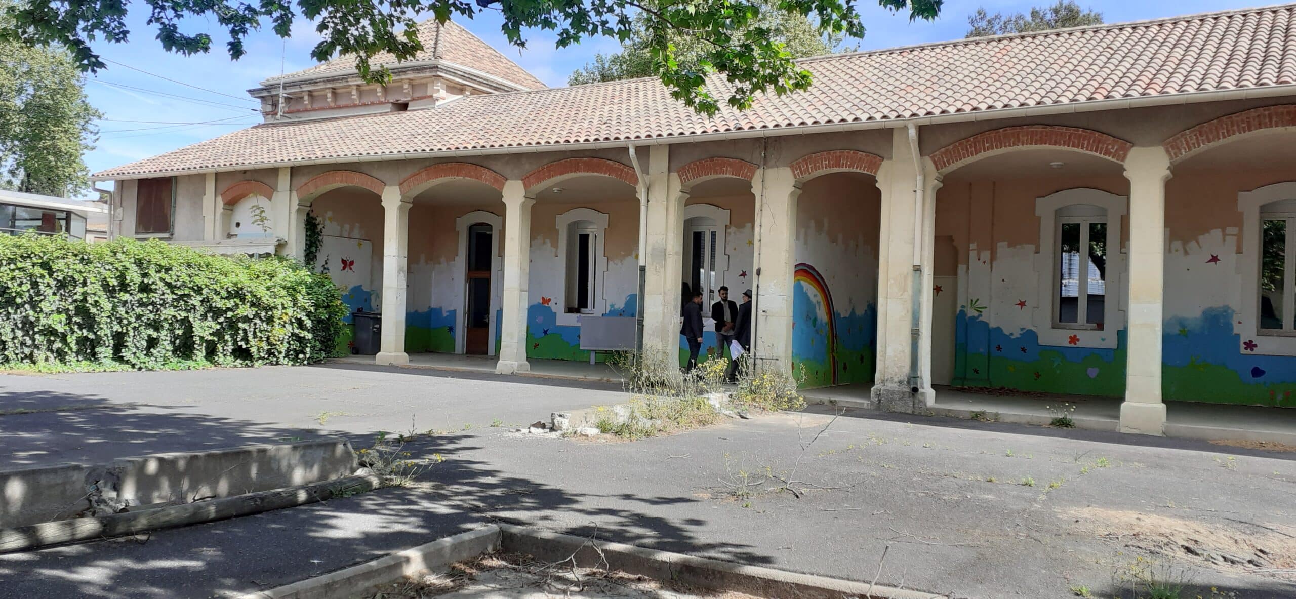 Bâtiment à arcades orné de fresques murales enfantines, avec trois personnes discutant sous le préau.
