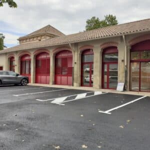 Bâtiment à arcades aux volets rouges et parking goudronné avec voiture grise garée devant.