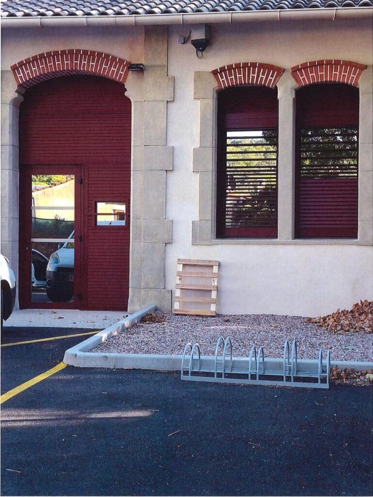 Bâtiment avec Porte Rouge et Rack à Vélos | Décor Urbain Porte et volets rouges d'un bâtiment avec rack à vélos, palette en bois et arches en briques rouges décoratives.