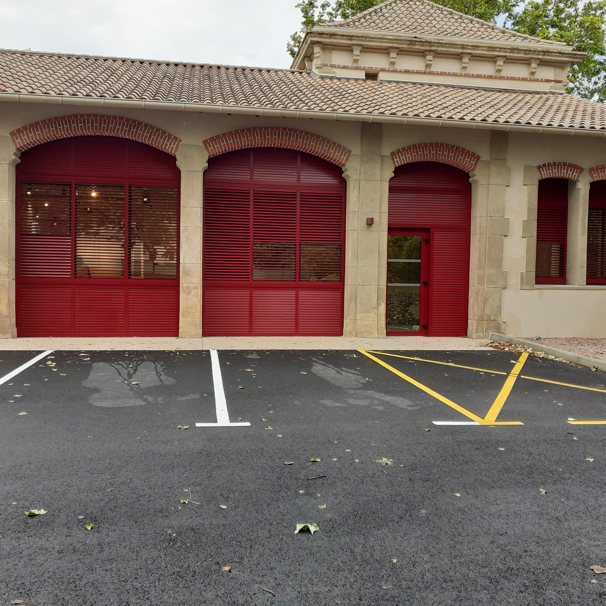 Bâtiment avec volets rouges à lattes, arches en brique et parking bitumé avec marquage jaune et blanc.