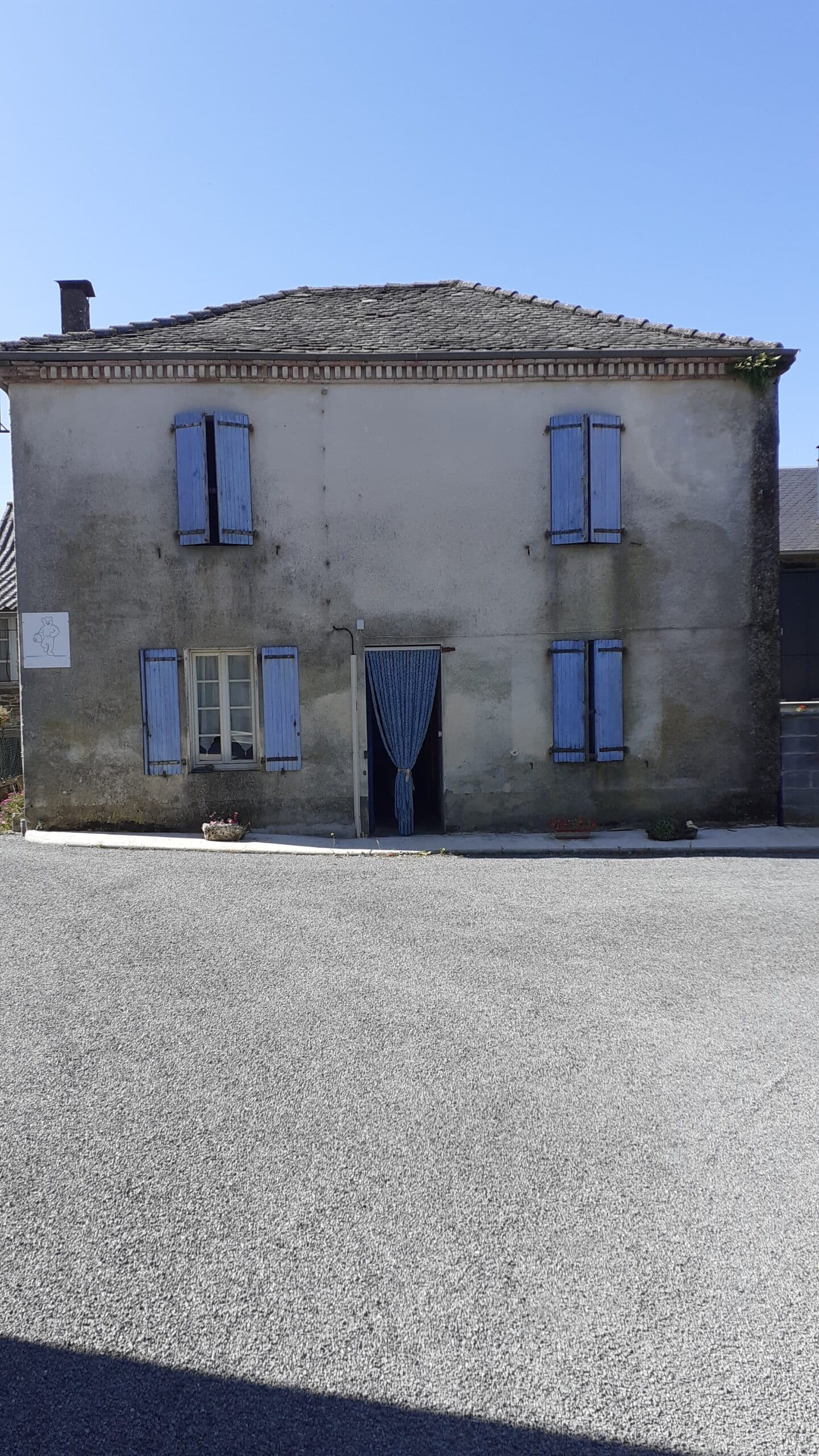 Maison ancienne avec volets bleus, rideau de porte et logo d'ours blanc sur une façade grise en crépi par temps clair.