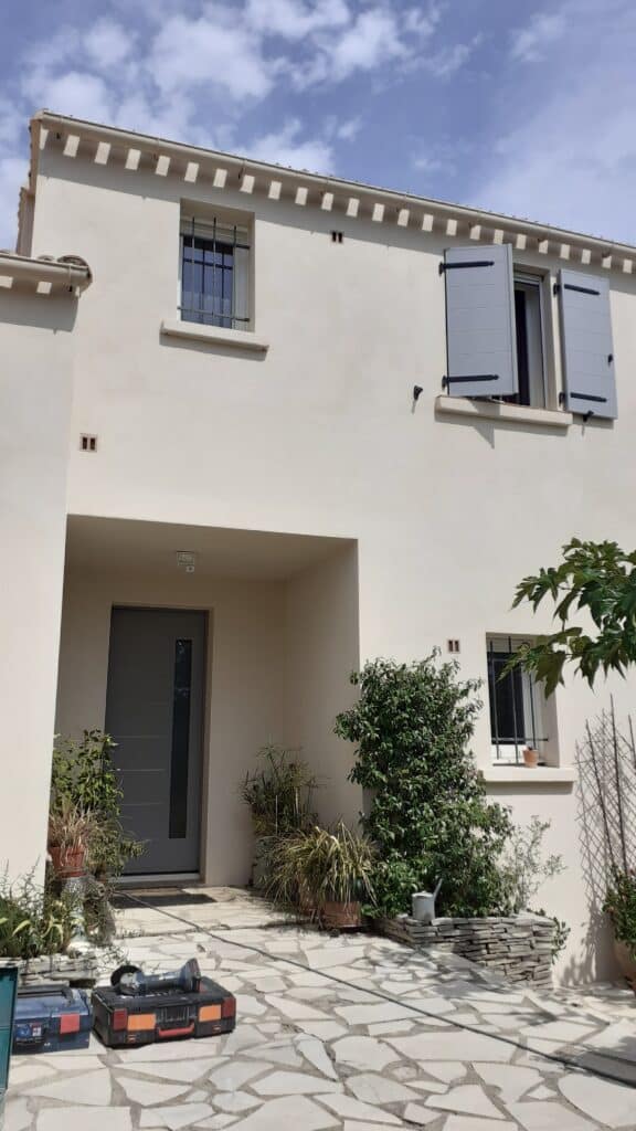 Maison moderne beige avec menuiseries grises et terrasse Maison moderne à façade beige avec porte grise, plantes en pots et outils sur une terrasse en pierre naturelle.