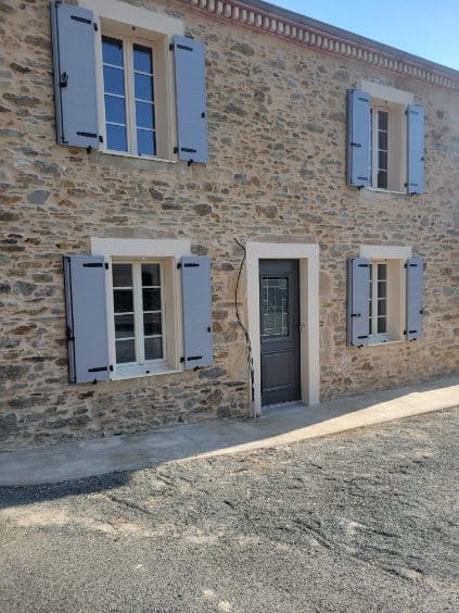 Maison en pierre traditionnelle avec ses volets bleus Façade de maison en pierre avec volets bleu clair, encadrements clairs et porte d'entrée gris foncé.