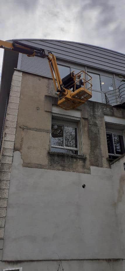 Nacelle élévatrice jaune et ouvrier effectuant des travaux de rénovation sur la façade d'un immeuble.