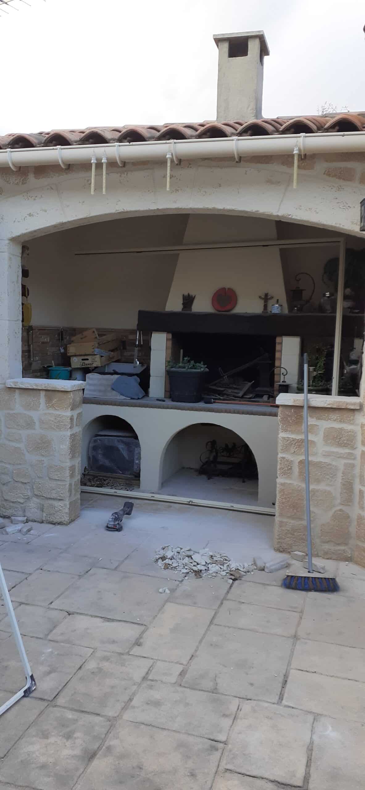 Cuisine d'été en pierre avec barbecue en cours de rénovation, débris au sol, meuleuse et balai sur un patio carrelé.