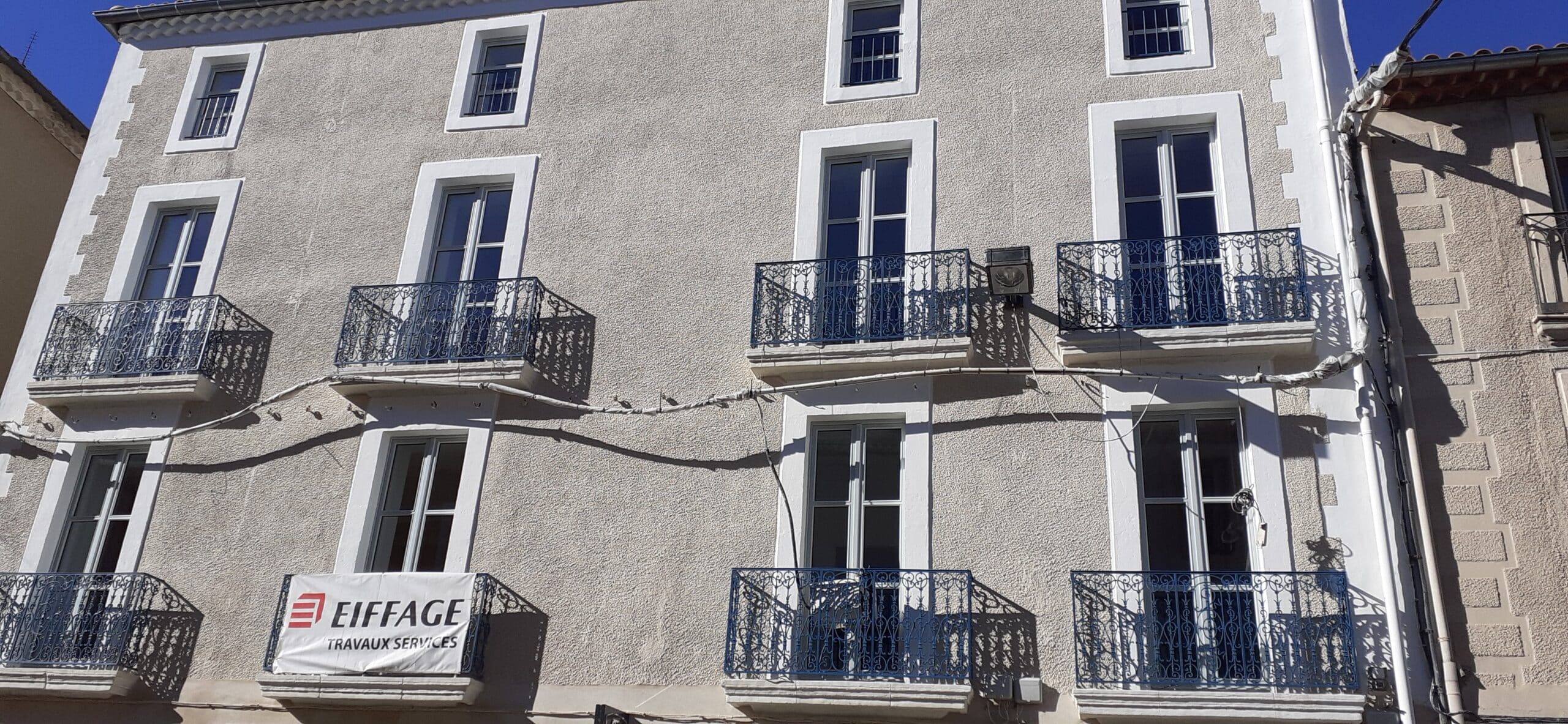 Rénovation de Façade par Eiffage Travaux Services Façade d'un immeuble gris avec balcons en fer forgé bleu et banderole Eiffage Travaux Services.