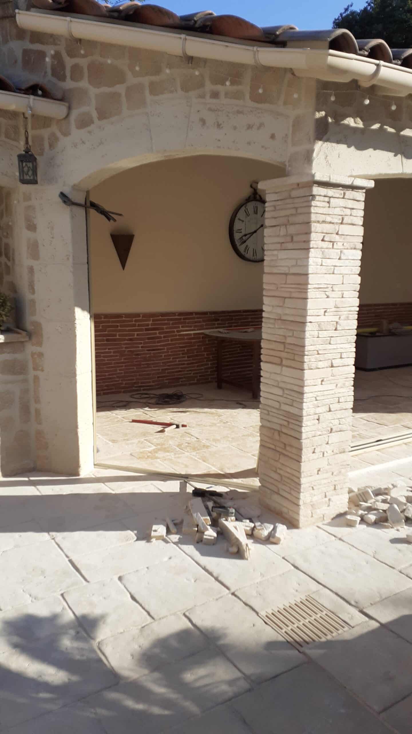 Terrasse en pierre naturelle en rénovation avec pilier en parement, horloge murale et débris de chantier au sol.
